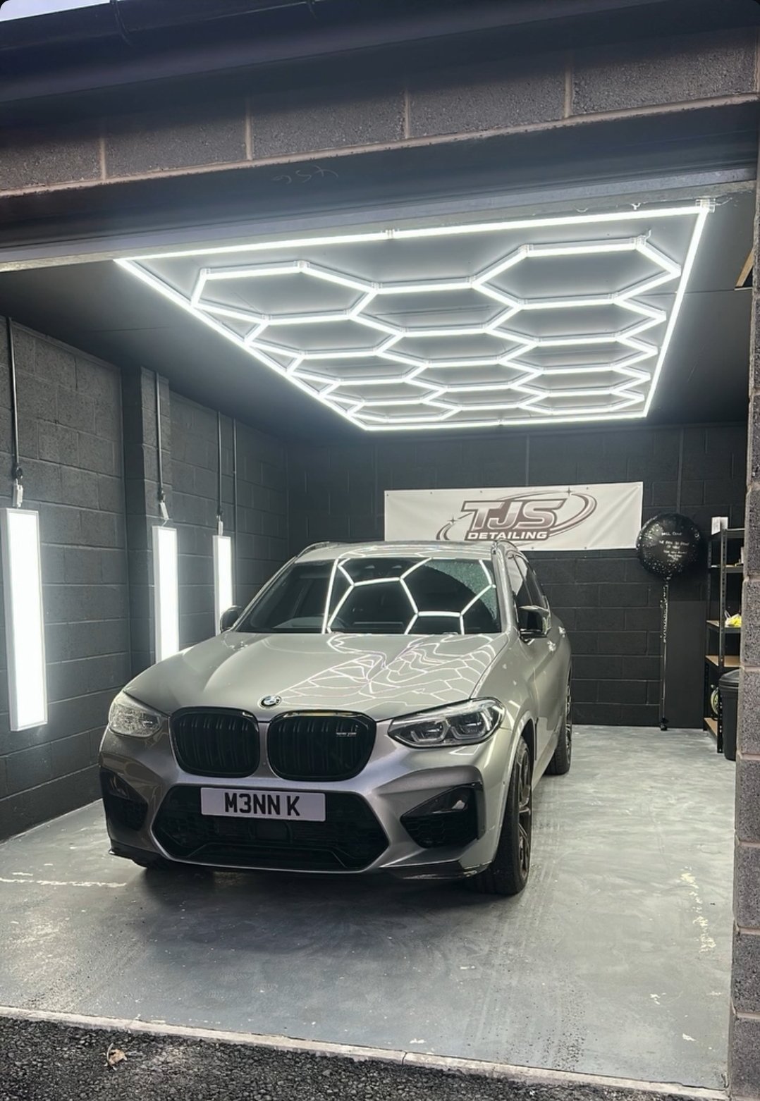 A BMW X3 M parked inside the new TJS Detailing unit, under white hexagon ceiling lights, with the TJS Detailing banner on the black block wall behind it